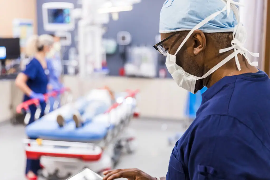 Male doctor reviewing patient data on tablet in busy hospital. Healthcare worker in scrubs.