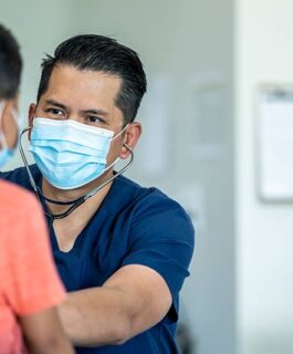 Medical professional wearing a mask and using a Stethoscope on a young patient.